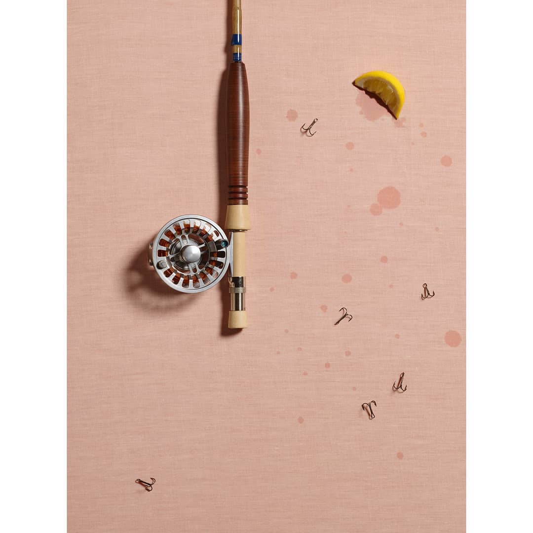 Fishing rod with fishing hooks and a lemon slice arranged on a blush pink surface with a minimalistic and clean aesthetic.