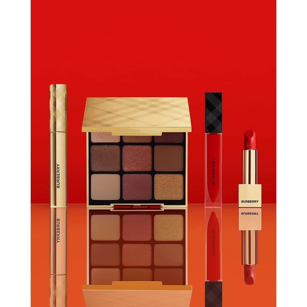 Burberry beauty product set against a red background includes eyeshadow palettes, lipstick, and lip gloss for a complete makeup collection.