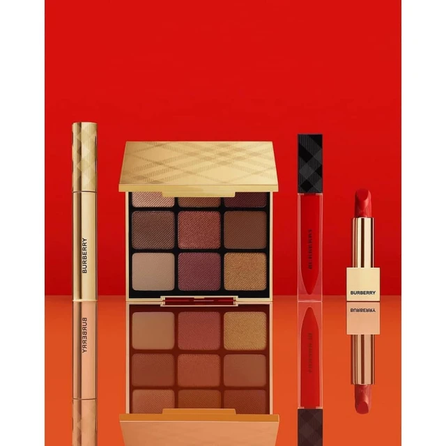 Burberry beauty product set against a red background includes eyeshadow palettes, lipstick, and lip gloss for a complete makeup collection.