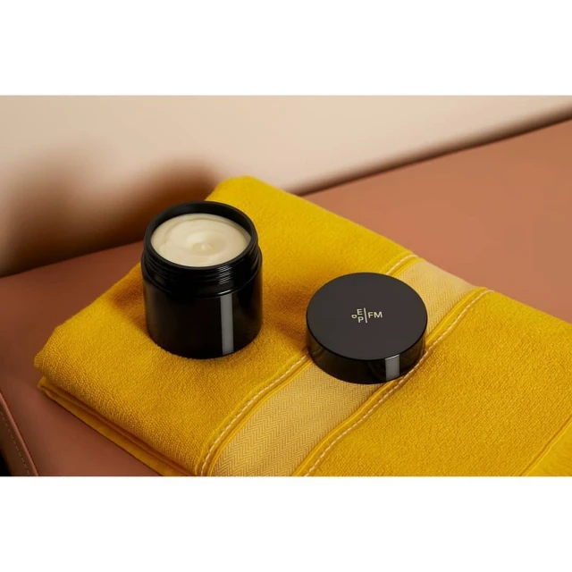 Cosmetic cream in a black container with a lid sits on a yellow striped towel against a two-tone background, product shot for beauty.