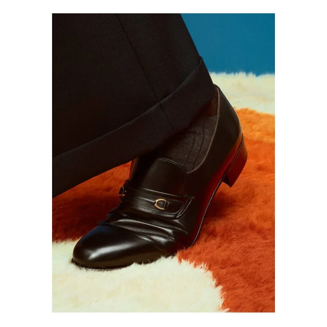 Close-up studio shot of a black leather loafer shoe with gold hardware, styled with a black trouser leg on a textured orange rug.