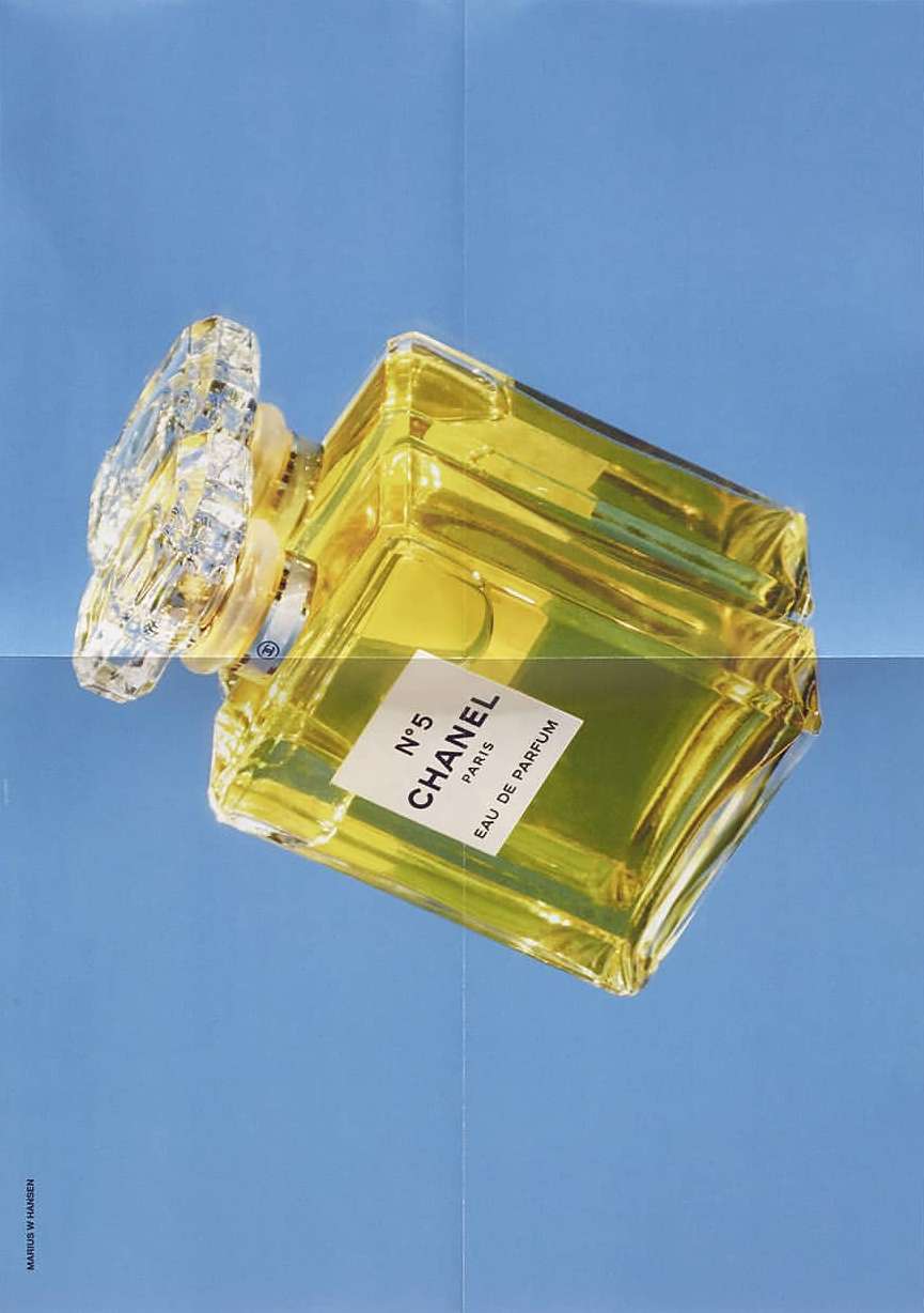 Chanel N°5 perfume bottle against a light blue background. The clear glass bottle has a yellow tint. The cap has a crystal design.