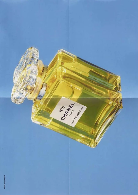 Chanel N°5 perfume bottle against a light blue background. The clear glass bottle has a yellow tint. The cap has a crystal design.