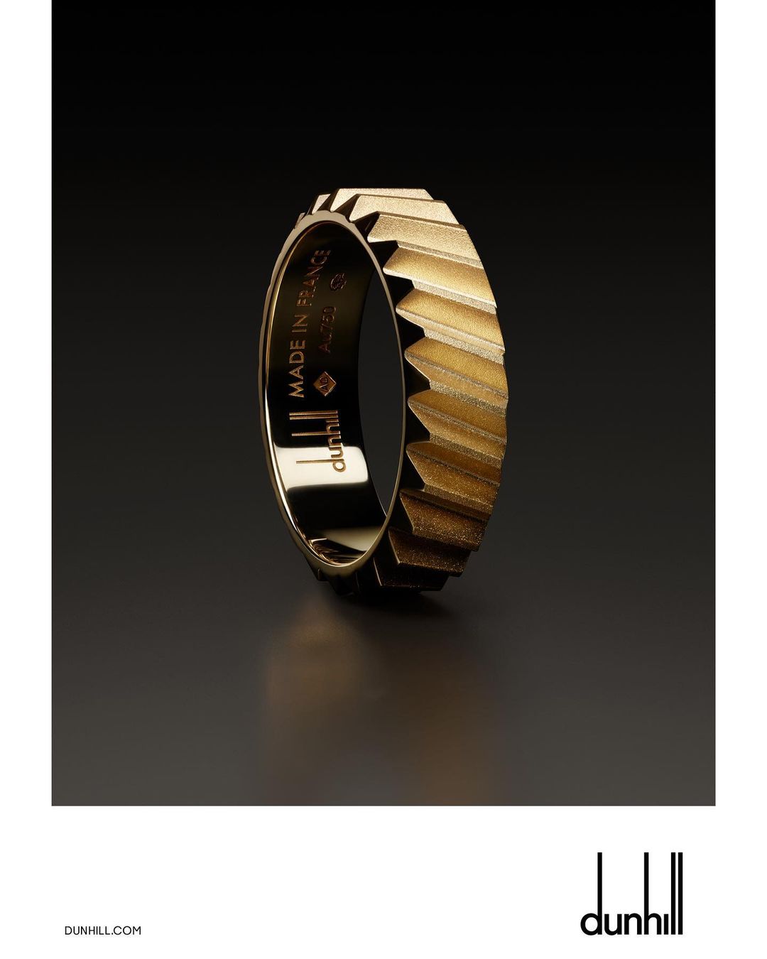 Close-up studio shot of a gold Dunhill ring with geometric details. The dark background contrasts with the reflective gold metal.