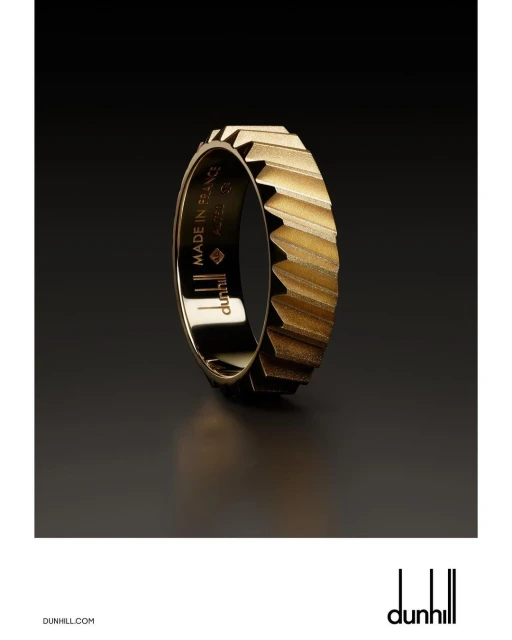 Close-up studio shot of a gold Dunhill ring with geometric details. The dark background contrasts with the reflective gold metal.