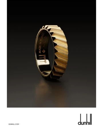 Close-up studio shot of a gold Dunhill ring with geometric details. The dark background contrasts with the reflective gold metal.