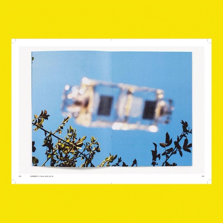 Open pages of a magazine with a blurred photograph of a vehicle against a blue sky, framed by branches and set on a bright yellow surface.