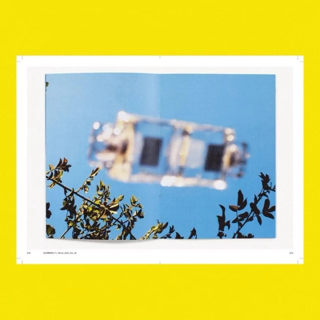 Open pages of a magazine with a blurred photograph of a vehicle against a blue sky, framed by branches and set on a bright yellow surface.