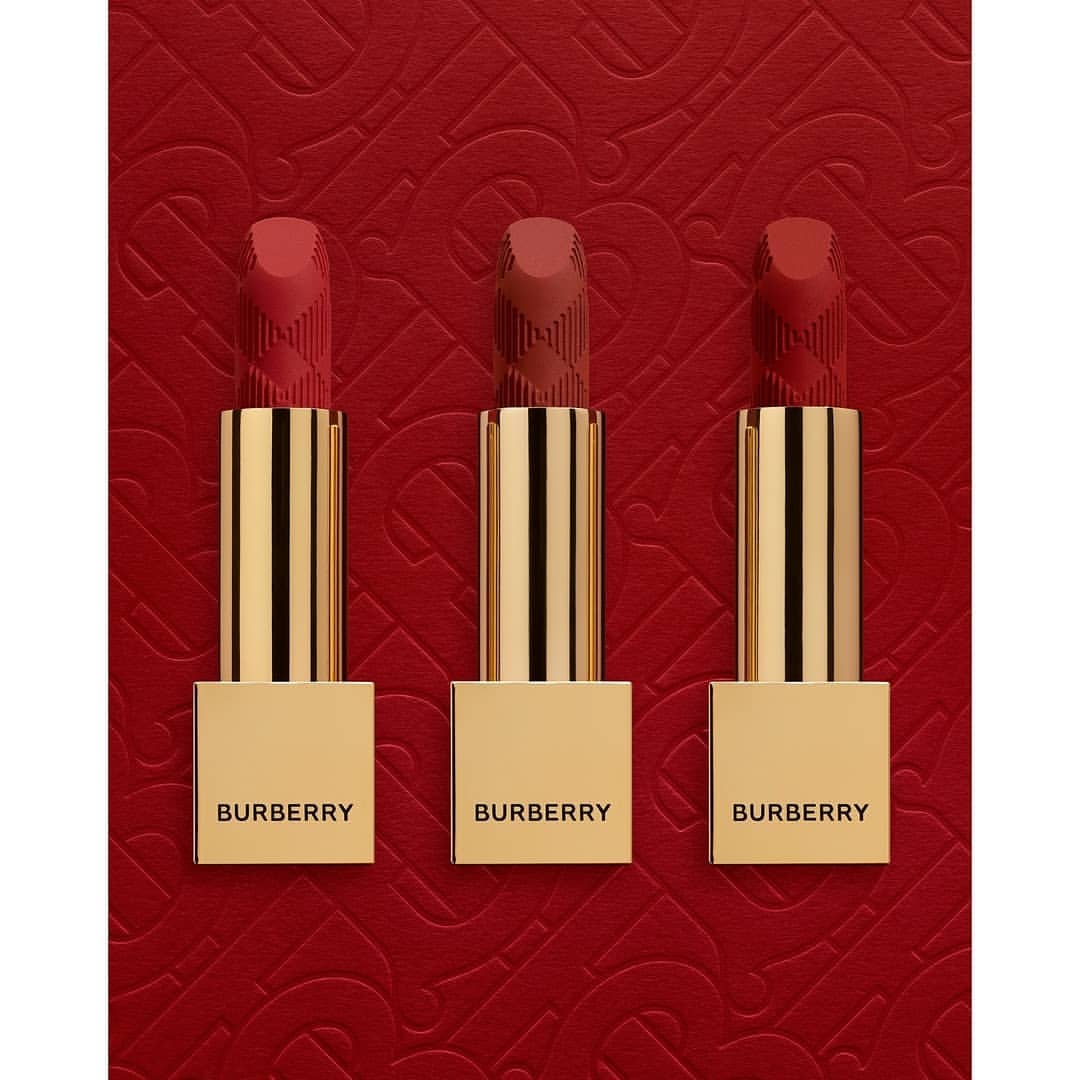 Three Burberry lipsticks in varying red hues stand upright with golden tubes against a textured red background with a subtle pattern.