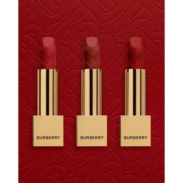 Three Burberry lipsticks in varying red hues stand upright with golden tubes against a textured red background with a subtle pattern.