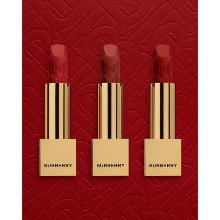 Three Burberry lipsticks in varying red hues stand upright with golden tubes against a textured red background with a subtle pattern.