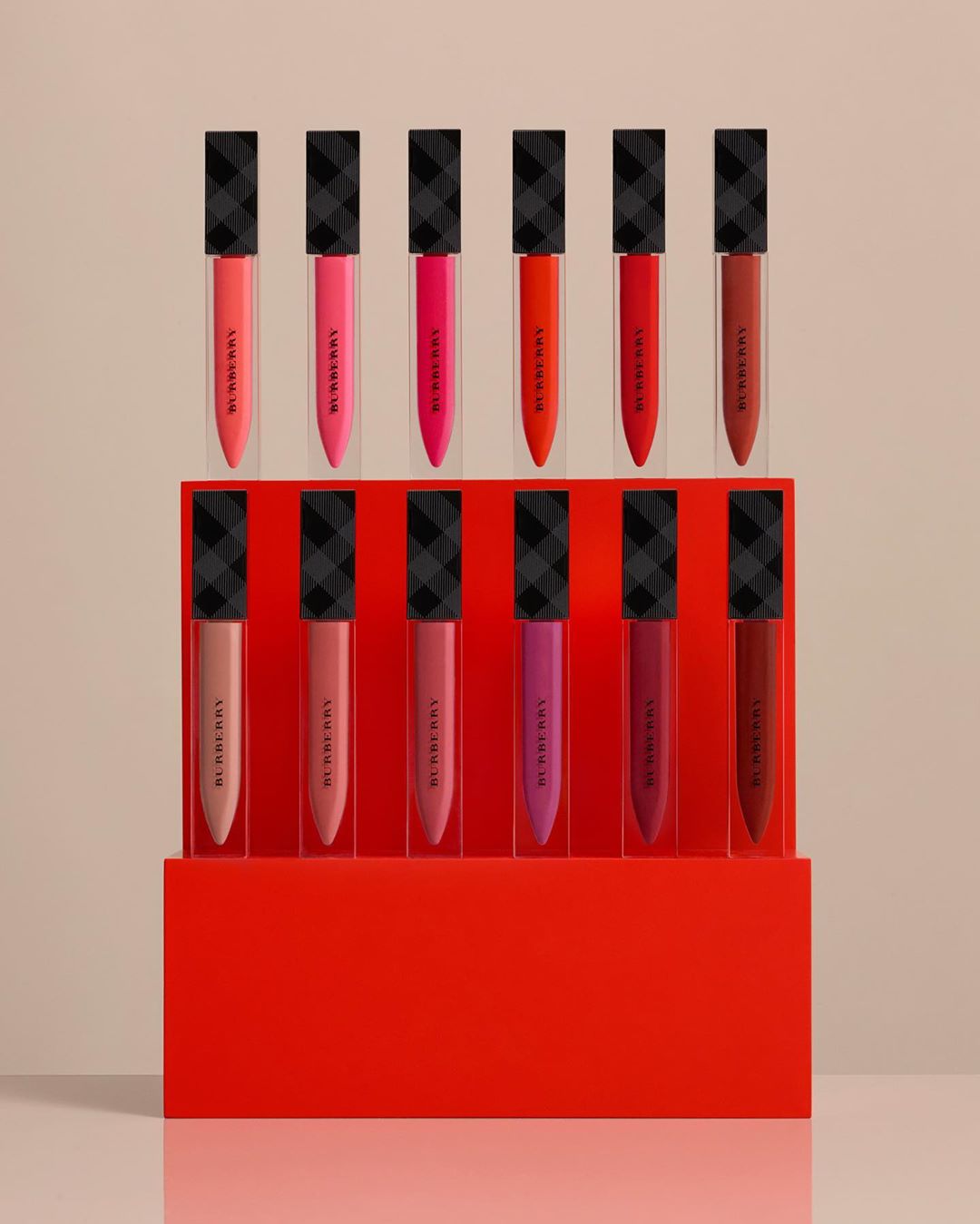 Burberry makeup product still-life: a collection of matte liquid lipsticks in a variety of red and neutral shades, arranged on a red display stand.