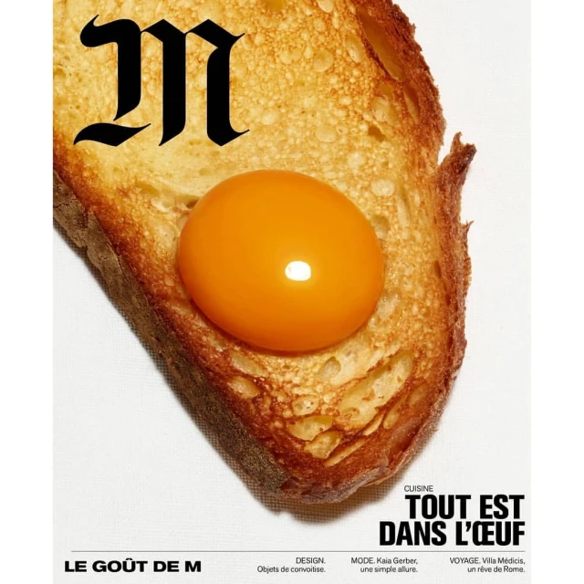 Overhead shot of a piece of golden toast topped with a bright orange egg yolk, next to a black stylized letter M on white.
