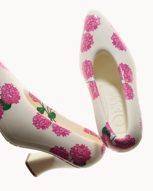 Two off-white high heel shoes are patterned with pink floral designs, set against a stark white backdrop, focusing on fashion footwear.