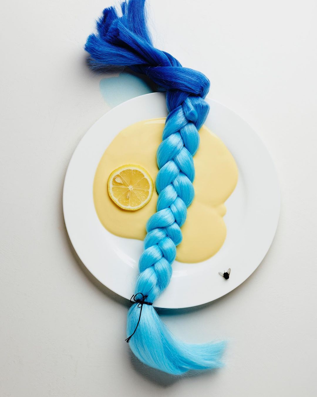Artistic studio shot of a blue braided wig on a white plate, paired with a lemon slice and light yellow liquid, creative composition.