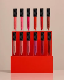 Twelve Burberry liquid lipstick tubes arranged in two rows on a bright red tiered display pedestal against a neutral studio background