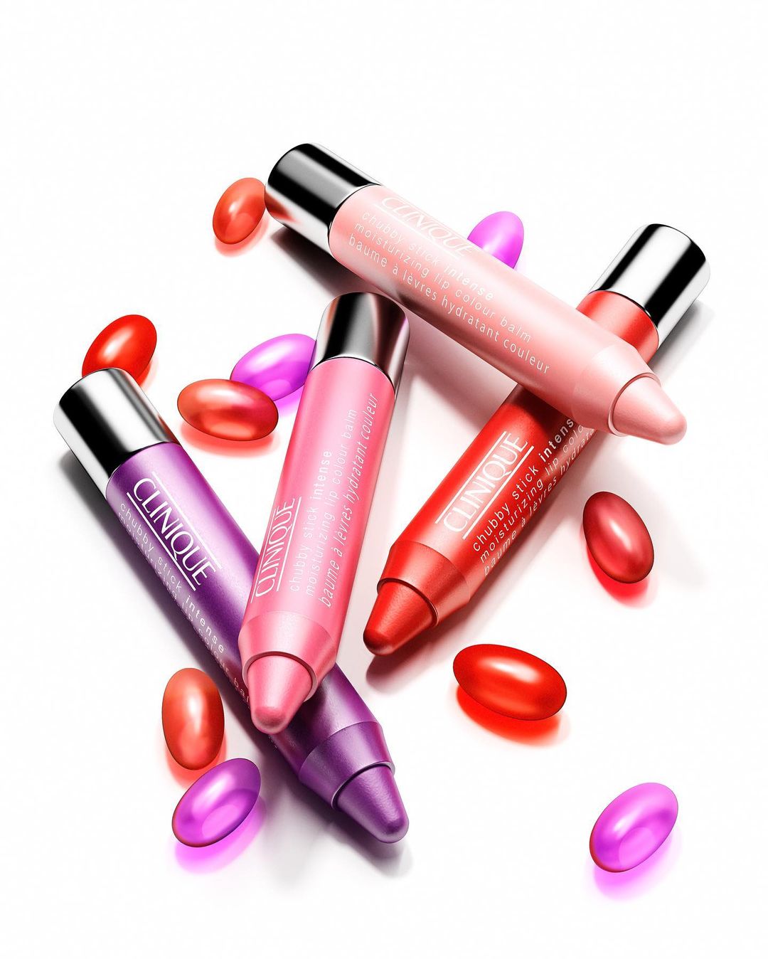 Clinique Chubby Stick Intense moisturizing lip color balms in pink, coral, and purple arranged with shiny red and purple capsules.