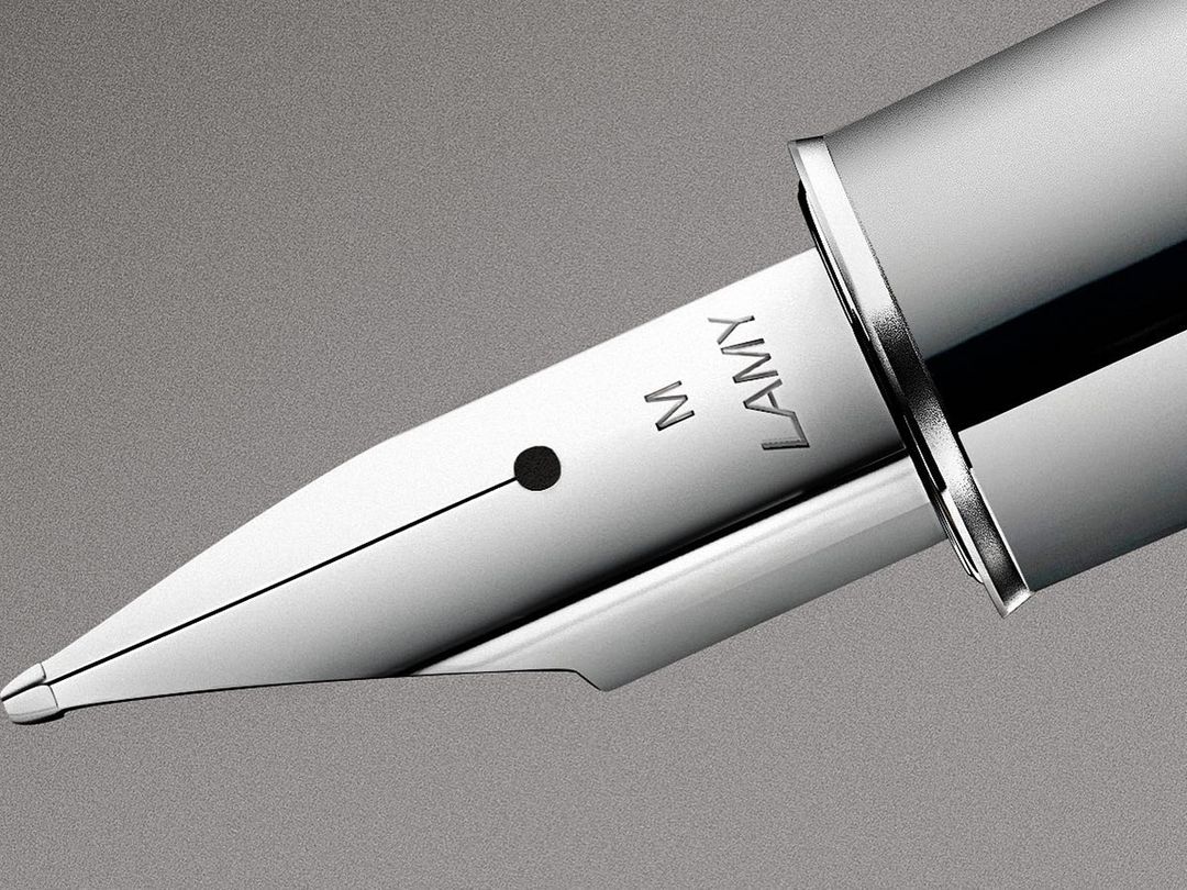 Close-up of a silver LAMY fountain pen nib with visible 'M' and 'LAMY' markings against a neutral gray background, showcasing its design and craftsmanship.