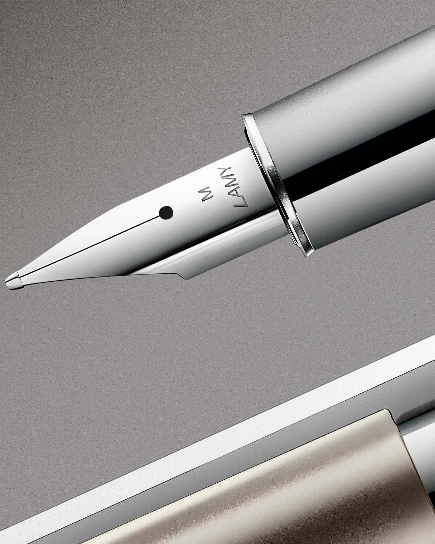 Close-up of a chrome and silver fountain pen featuring the brand 'Lamy' and a nib marked with the letter 'M' against a gray backdrop.