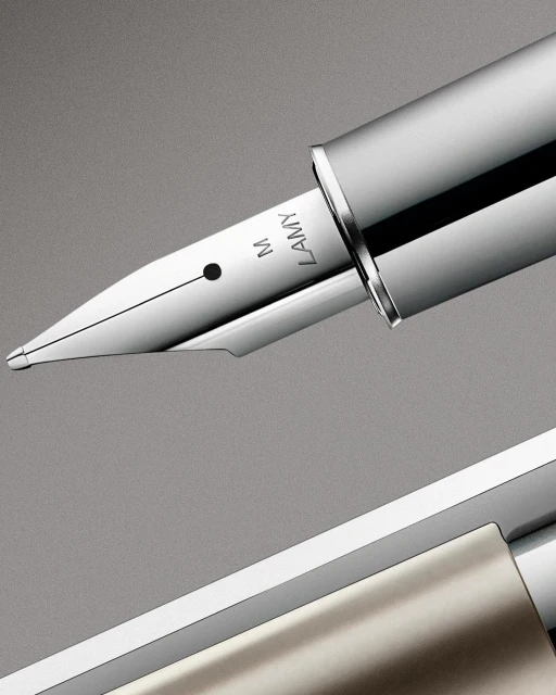 Close-up of a chrome and silver fountain pen featuring the brand 'Lamy' and a nib marked with the letter 'M' against a gray backdrop.