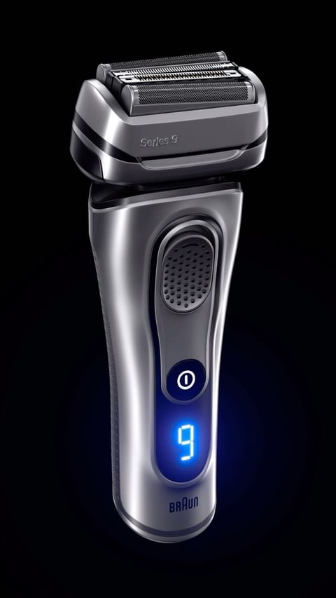 Close-up studio shot of a silver and black Braun Series 9 electric shaver on a black background with blue illumination and digital display.