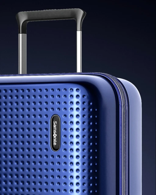 Close-up of a blue Samsonite suitcase showing the textured surface, logo, telescoping handle, and metallic frame details against a dark background.