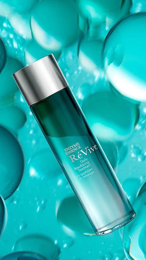 ReVive Enzyme Essence bottle on a blue surface surrounded by translucent blue spheres. Silver top and branding on the label.