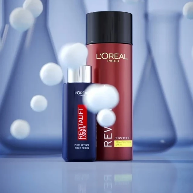 Two L'Oreal product bottles, a dark blue night serum and a red sunscreen, are staged in a skincare product shot with laboratory glassware.