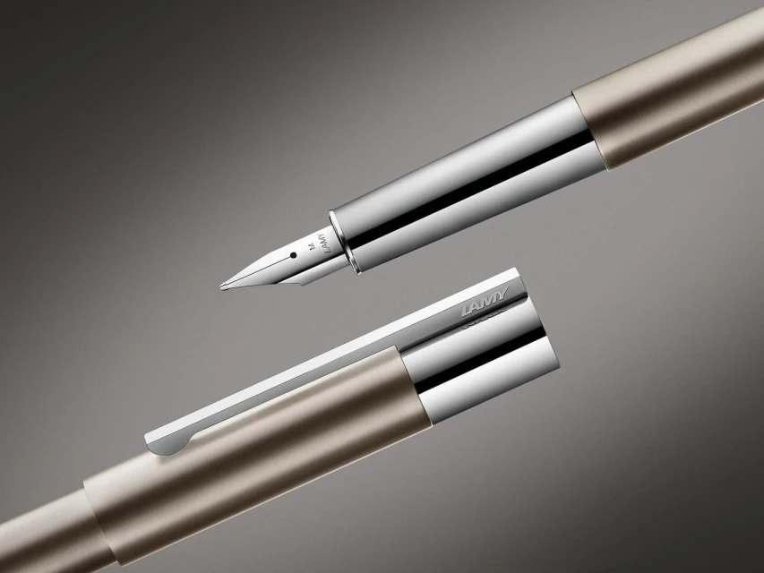 Close-up product shot of a Lamy fountain pen with a metallic gold barrel, silver nib, and a detached cap against a gradient grey background.