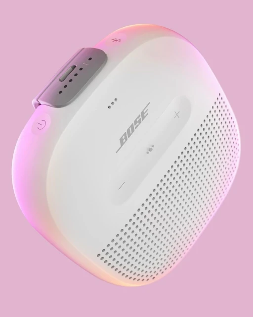 Close-up studio shot of a white Bose Bluetooth speaker against a pink background with colorful rim lighting effect emphasizing its features.
