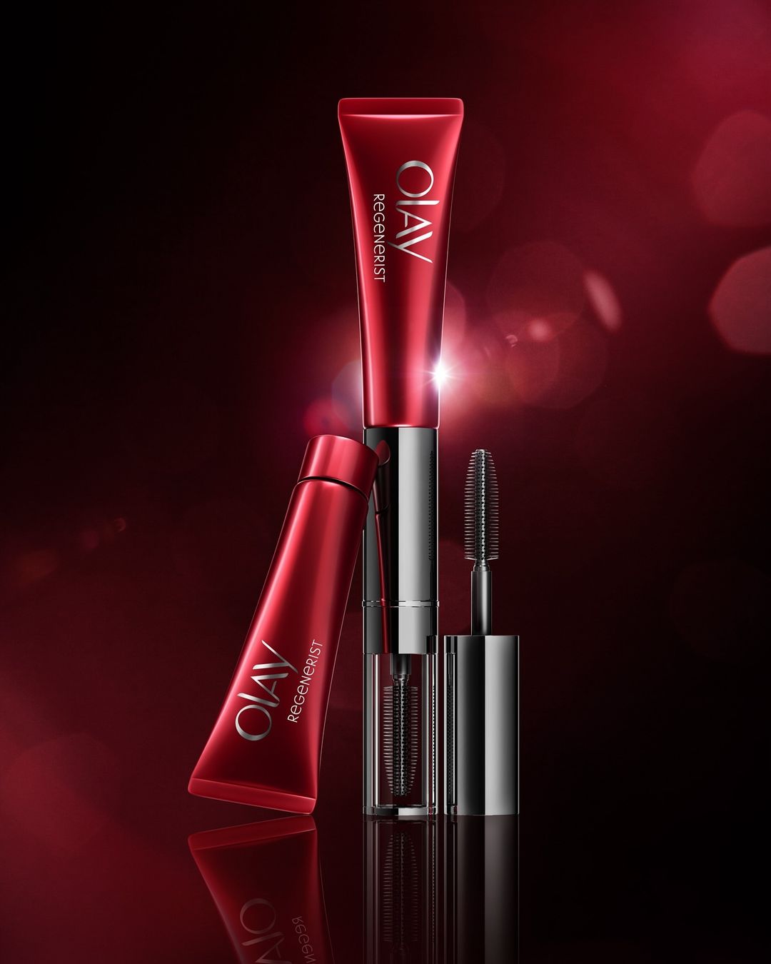 Red and silver Olav Regenerist beauty product tubes and mascara wand on a dark red background with blurred bokeh effects.
