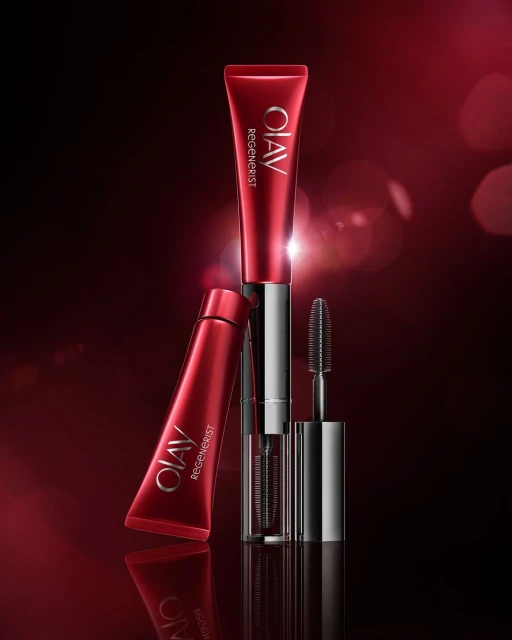 Red and silver Olav Regenerist beauty product tubes and mascara wand on a dark red background with blurred bokeh effects.