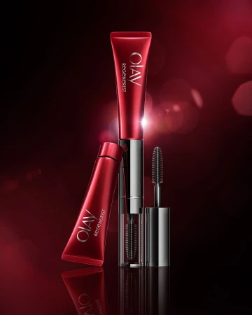 Red and silver Olav Regenerist beauty product tubes and mascara wand on a dark red background with blurred bokeh effects.