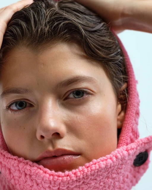 Close-up portrait of a young fair-skinned woman with blue eyes wearing a pink hooded jacket in a studio setting, hands near her head.