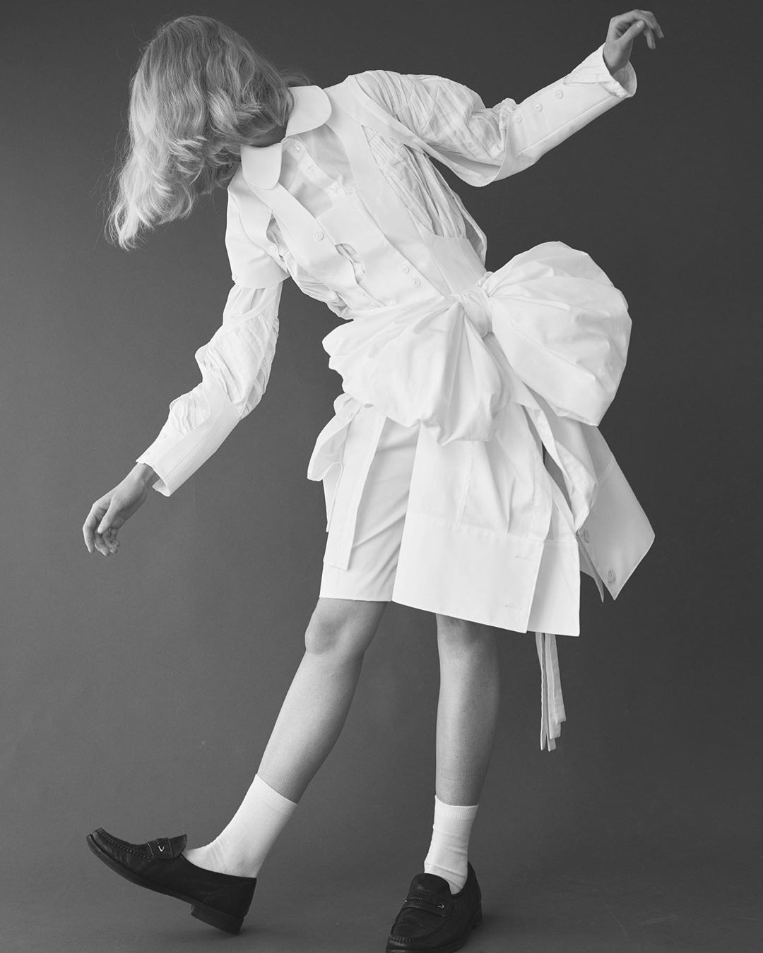 Person with blond hair poses in a white deconstructed shirt dress with oversized bow, white socks, and black loafers against gray background.