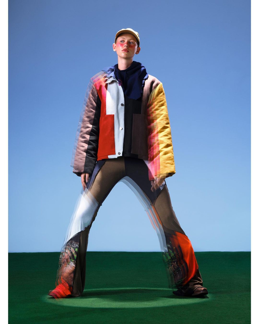 Fashion shot of a person wearing a colorful patchwork jacket and patterned pants, posing in front of a clear blue sky and green field.