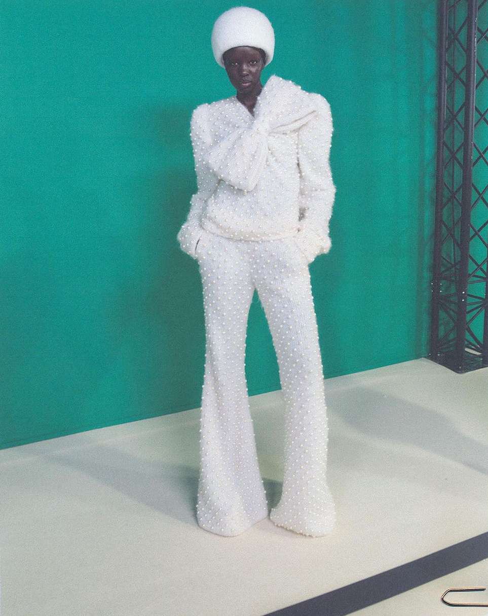 A slender African woman in a white, textured pantsuit and matching hat poses against a green backdrop in a full, eye-level shot.