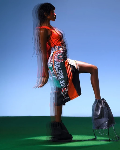 Adult female fashion model poses in an orange tank top and patchwork NBA basketball jersey dress with drawstring bag shoes against a blue sky.