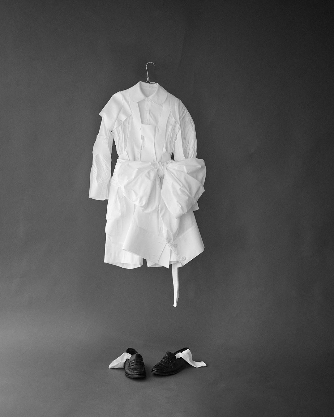Fashion shot of a white dress shirt on a hanger above black dress shoes with white fabric accents against a gray studio background.