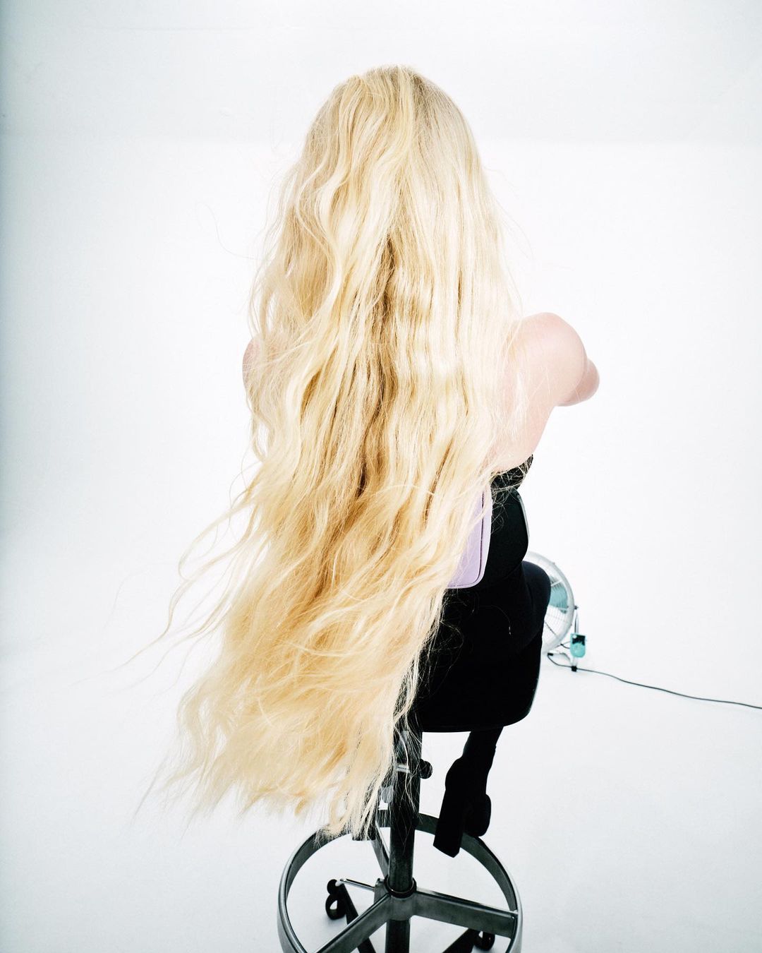 Back view of a woman with long blonde wavy hair, sitting on a rolling stool in a white studio, showcasing hair and styling for beauty.
