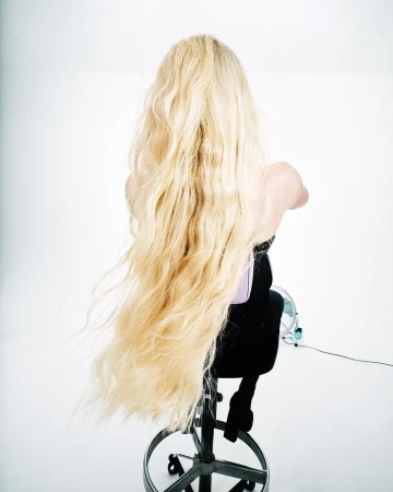 Back view of a woman with long blonde wavy hair, sitting on a rolling stool in a white studio, showcasing hair and styling for beauty.