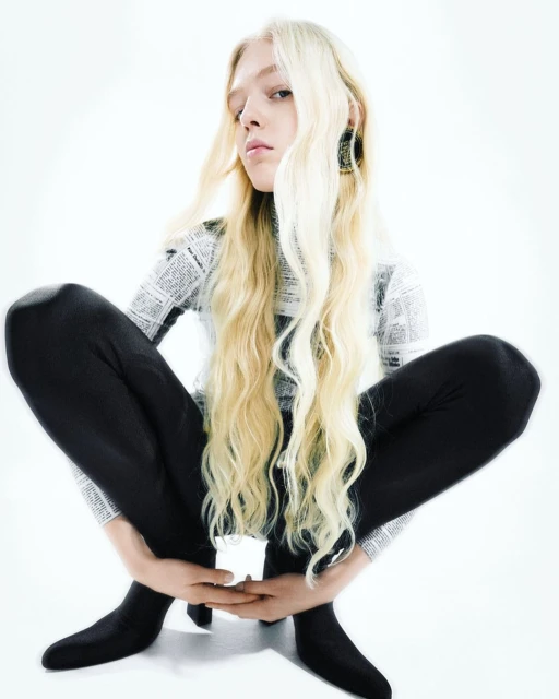 A slender adult woman with blonde wavy hair sits in a squatting position in a studio wearing a newspaper-print long-sleeve shirt and black leggings.