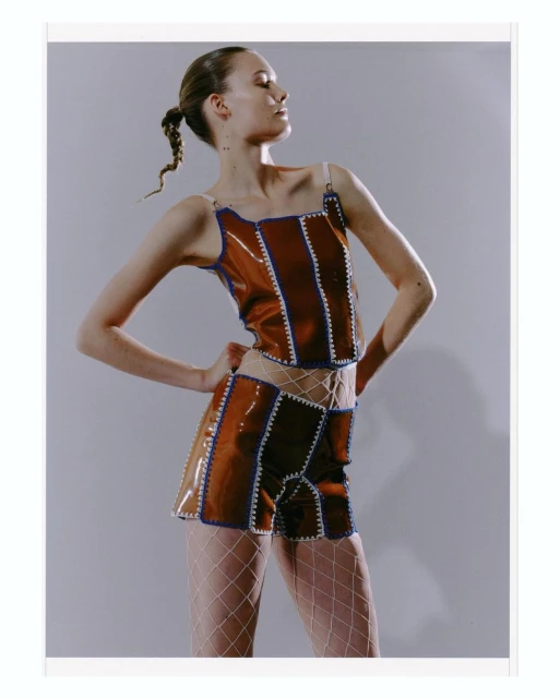 A model poses in a brown two-piece outfit with white fishnet stockings, against a neutral backdrop in a studio fashion shot.