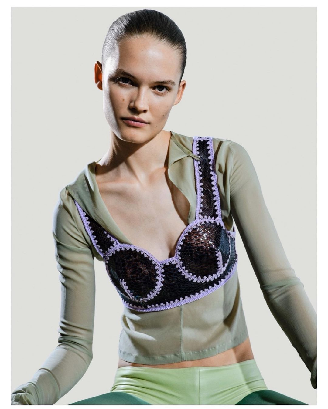 Fashion photo of a woman with slicked-back hair wearing a unique sequined and crochet-trimmed top, styled with a sheer blouse and pants.