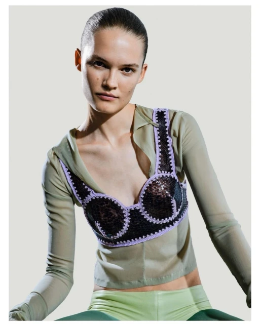Fashion photo of a woman with slicked-back hair wearing a unique sequined and crochet-trimmed top, styled with a sheer blouse and pants.