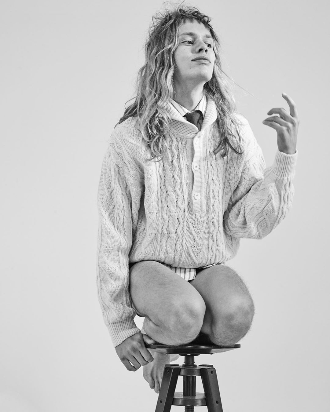 Monochrome portrait of a fair-skinned, long-haired person kneeling on a stool, wearing a white sweater and tie against a bright background.