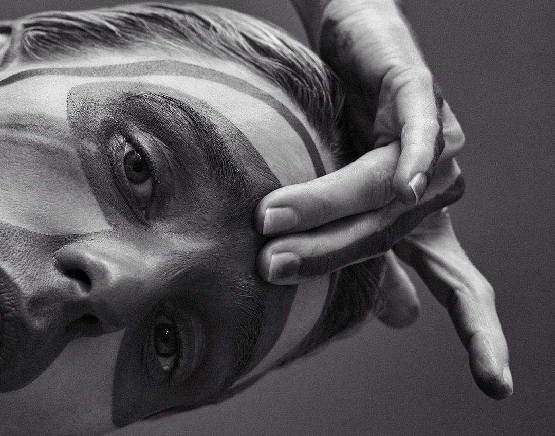 Monochrome portrait of a person with painted facial stripes and a hand partially covering their face in an artistic composition.