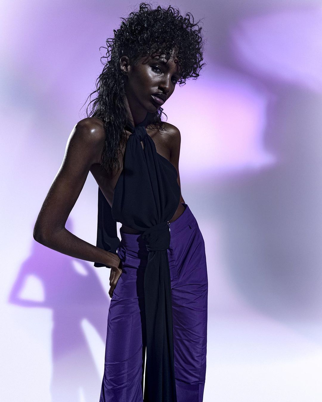Fashion shot of an adult female model with wet curly hair posing in a black top and purple pants against a mottled purple background.