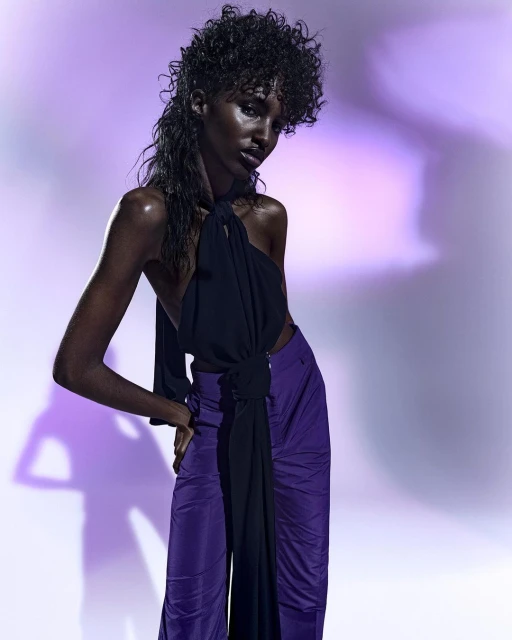Fashion shot of an adult female model with wet curly hair posing in a black top and purple pants against a mottled purple background.