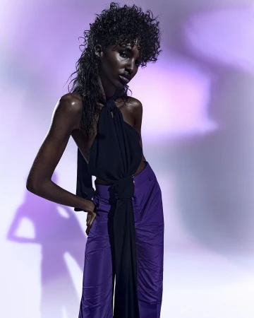Fashion shot of an adult female model with wet curly hair posing in a black top and purple pants against a mottled purple background.